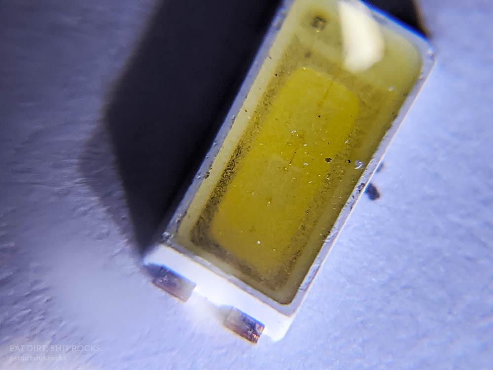 One of the LEDs on a note card. Beneath the yellow phosphor material, you can see the outlines of two square LED chips (this is a dual-chip LED) connected by fine bond wires.